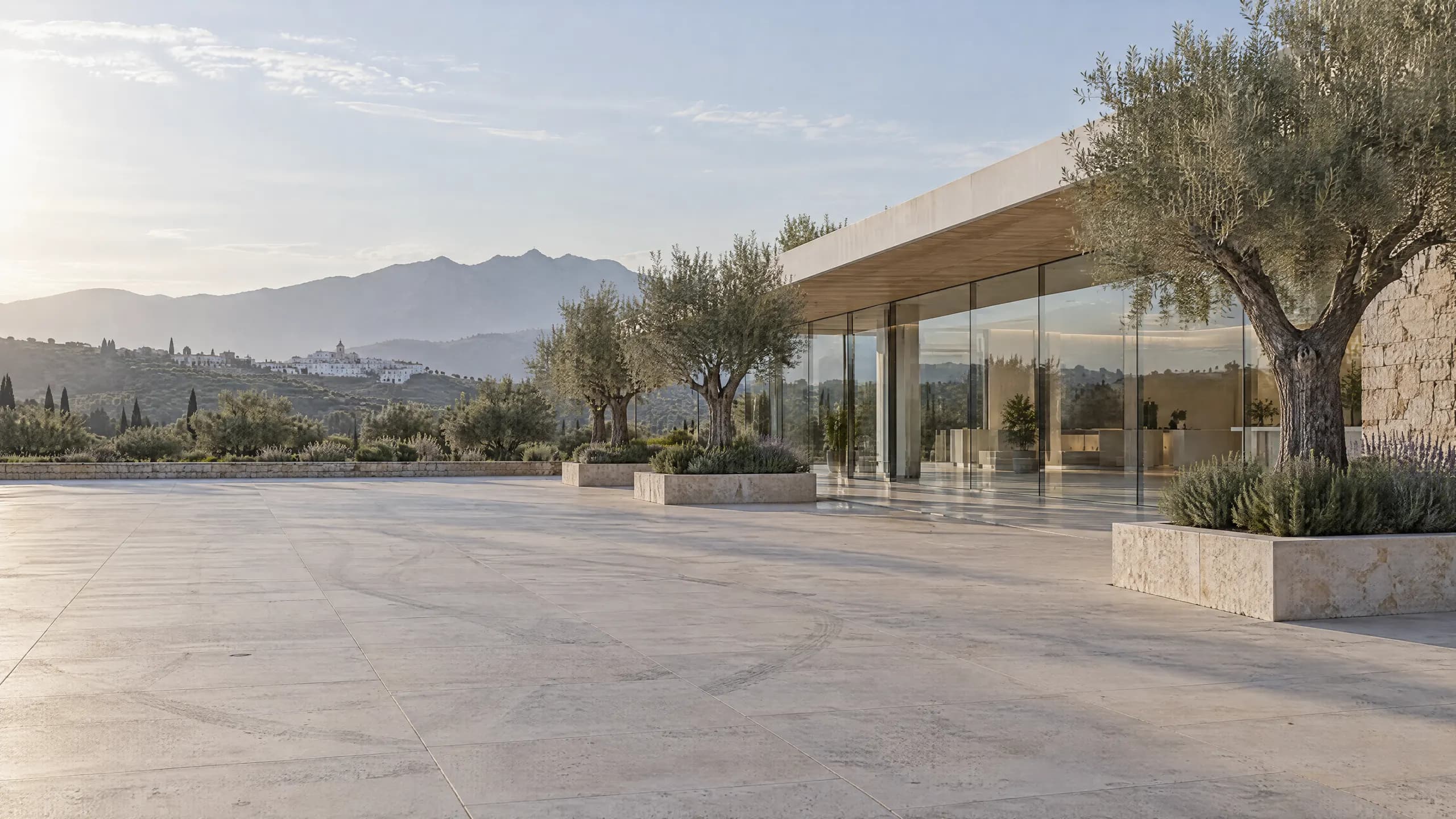 Explanada vacía de piedra caliza frente a un concesionario contemporáneo de cristal y piedra, con olivos en jardineras, a primera hora de la mañana.