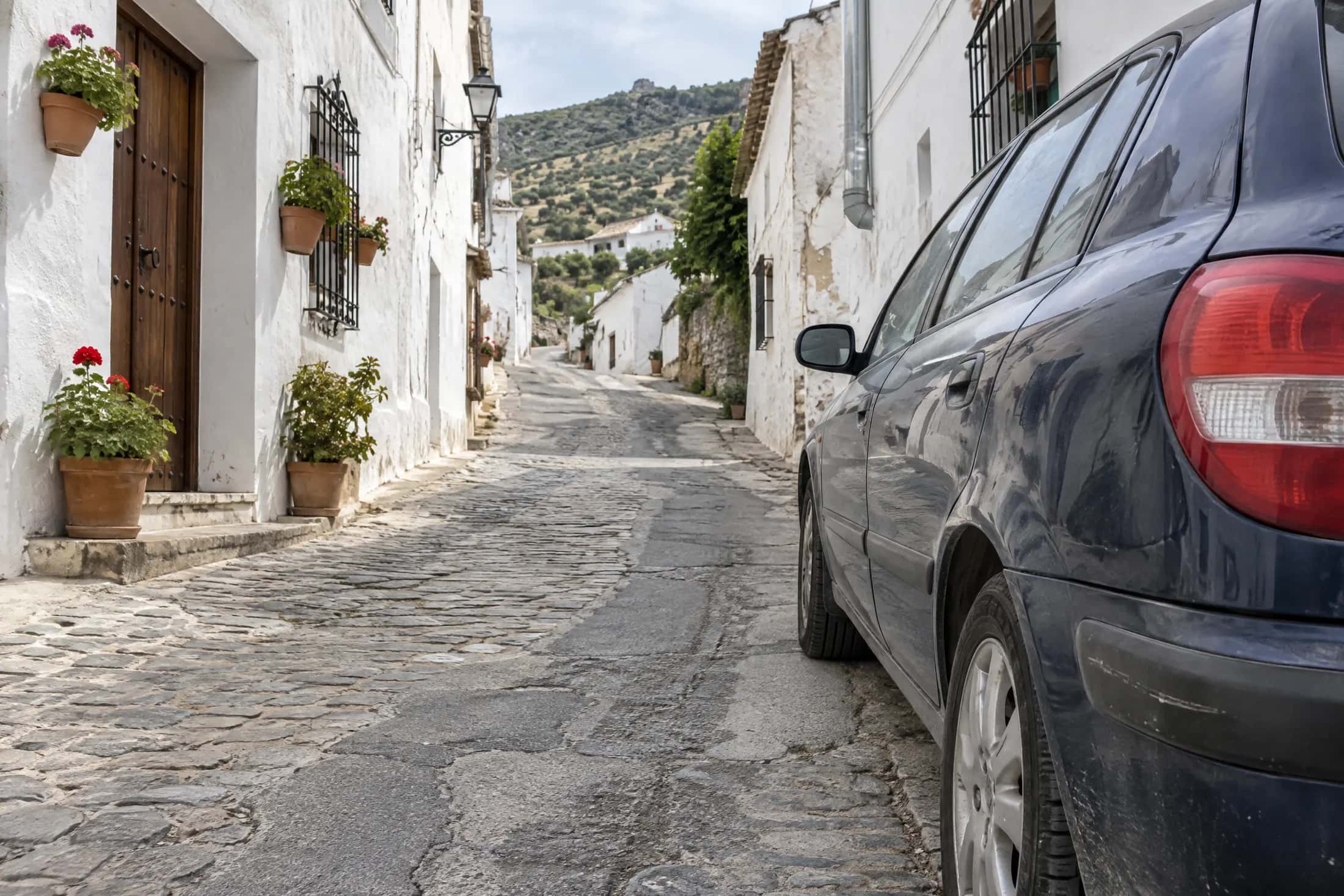 Calle empedrada en cuesta de un pueblo blanco de Málaga, con macetas de geranios y un coche azul aparcado junto a la fachada.