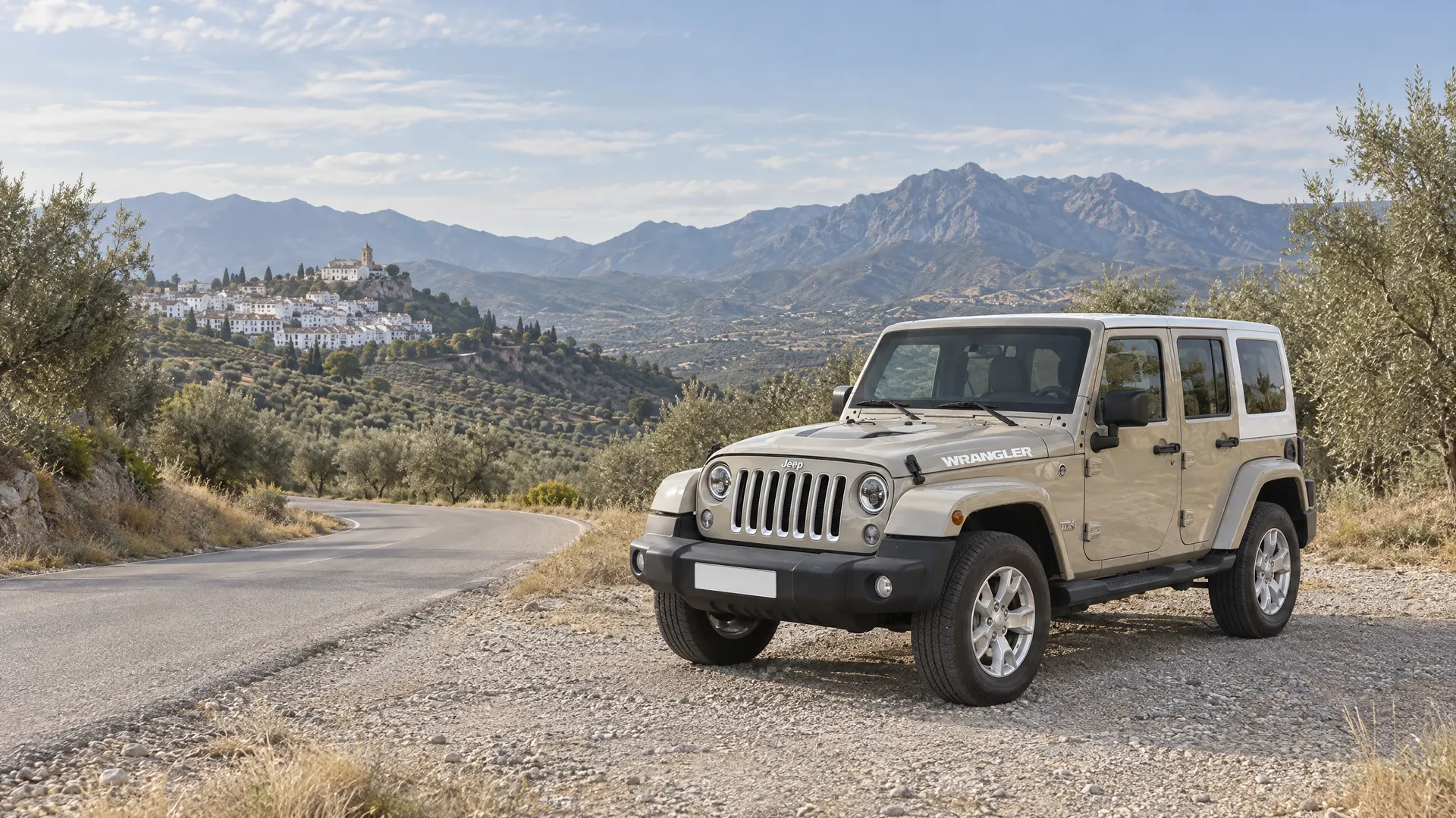 Jeep Wrangler color arena con techo duro blanco aparcado en el arcén de grava de una carretera de curvas entre olivos en la sierra de Málaga, con un pueblo blanco y la sierra al fondo.